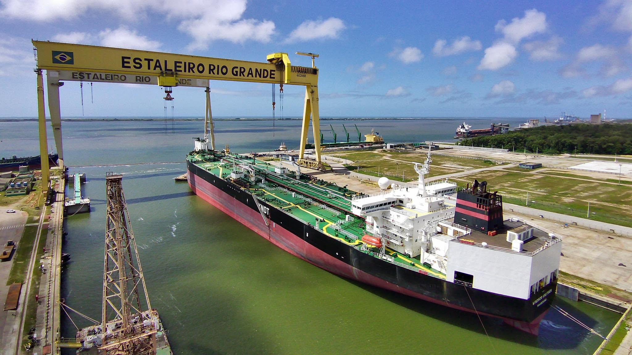 A large tanker vessel under construction in a shipyard, with cranes and workers visible.